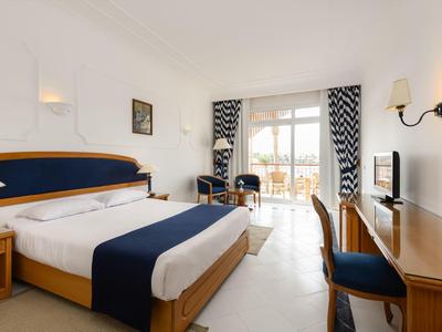 Lichte hotelkamer met tweepersoonsbed, bureau, stoelen en balkon met uitzicht naar buiten.