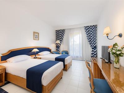 Lichte hotelkamer met twee bedden, bureau, bank en balkon met gordijnen.