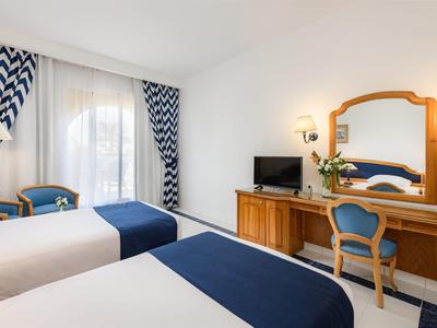 Lichte hotelkamer met twee bedden, blauw-witte gordijnen en houten bureau met spiegel.