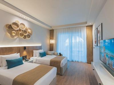 Modern hotelkamer met twee bedden, decoratieve wand, tv en groot raam met gordijnen.