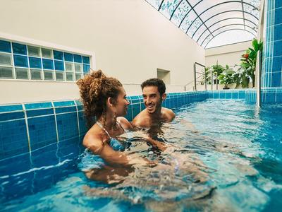 Twee personen ontspannen en praten in het blauwe binnenzwembad onder natuurlijk licht.