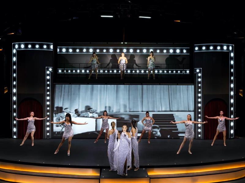 Podiumvoorstelling met elf dansers in witte kostuums en verlicht podiumontwerp.