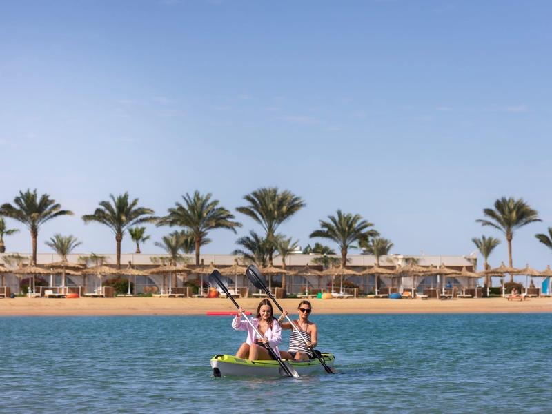 Twee personen peddelen in een kajak op kalm water met palmbomen en strand op de achtergrond.