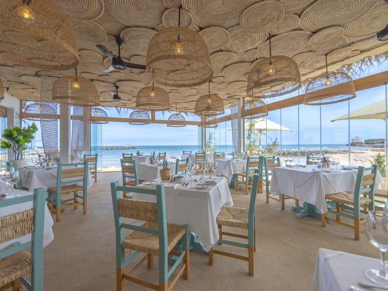 Licht restaurant met gedekte tafels en rieten lampen, uitzicht op zee en parasols buiten.