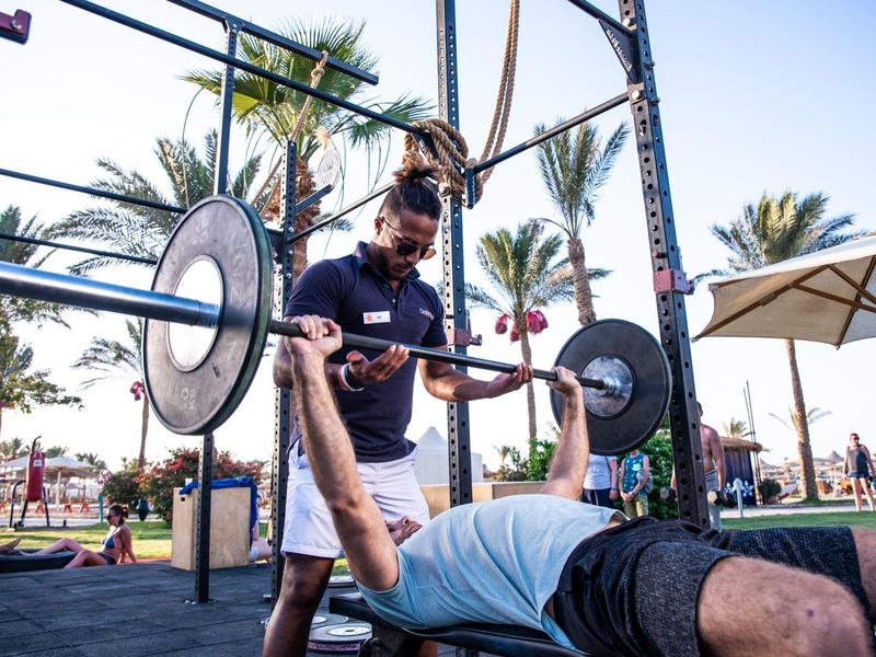 Man doet bankdruktraining buiten met trainer onder palmbomen en parasol.