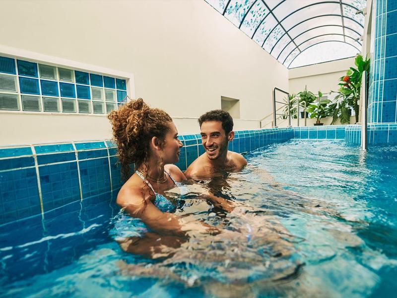 Twee personen ontspannen en praten in het blauwe binnenzwembad onder natuurlijk licht.