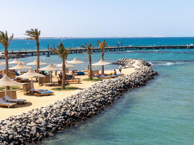Strandboulevard met palmbomen, parasols en ligstoelen aan helderblauwe zee.