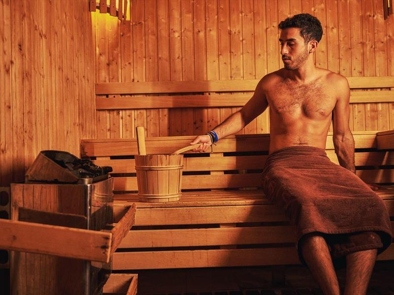 Man in sauna zit op houten bank naast emmer en schep.