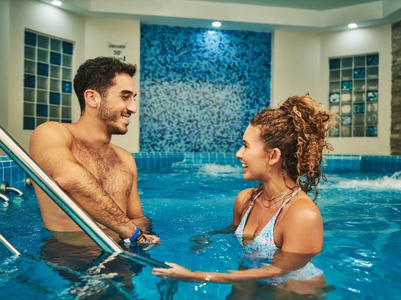 Een stel praat in een binnenzwembad van een hotel met een waterval op de achtergrond.