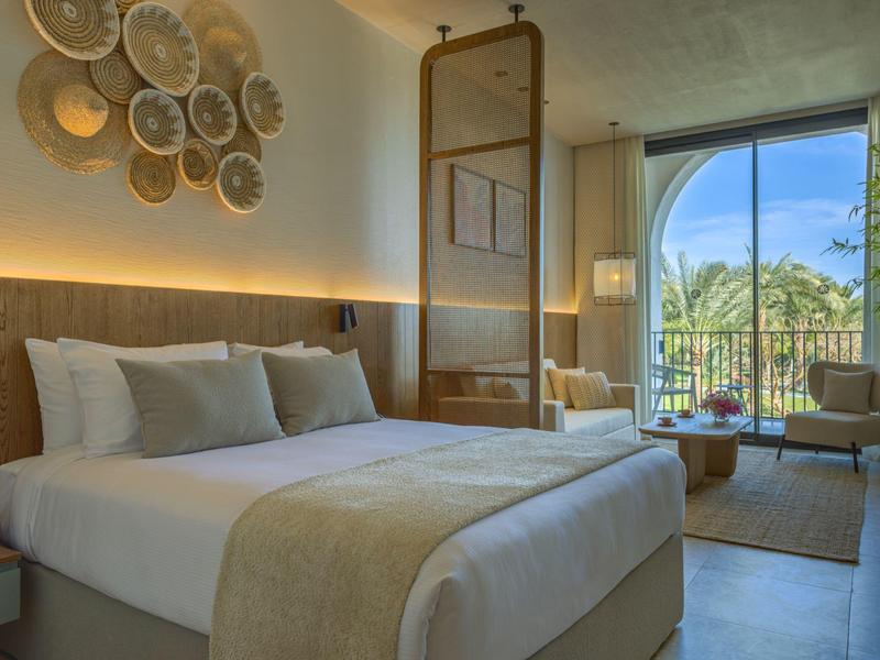 Modern hotelkamer met groot bed, zithoek en balkon met uitzicht op palmbomen.