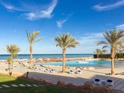 Strandpromenade mit Palmen, Liegestühlen und Blick auf das blaue Meer bei klarem Himmel.