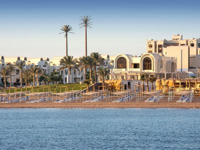 Strand mit Liegestühlen vor einem Hotel und Palmen bei Sonnenuntergang.