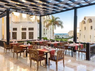 Offener Hotelrestaurantbereich mit Tischen, Stühlen und Blick auf Palmen und Meer.