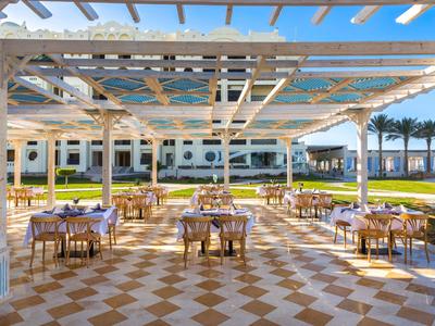 Freiluftrestaurant mit Holztischen und blauen Tischdecken unter Pergola mit Meerblick.
