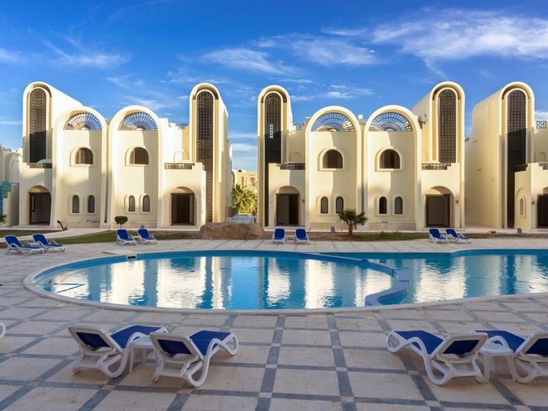 Nowoczesny kompleks hotelowy z białą elewacją i dużym basenem pod błękitnym niebem.