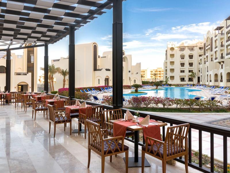 Zadaszony taras na zewnątrz z stołami i krzesłami obok basenu hotelowego i białych budynków.