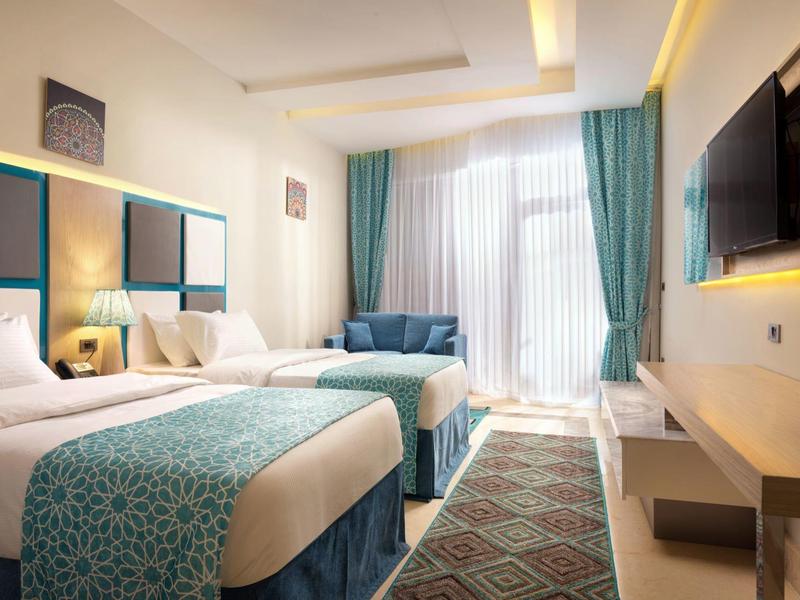 Jasny pokój hotelowy z dwoma łóżkami, niebiesko-zielonymi zasłonami i nowoczesnym telewizorem.