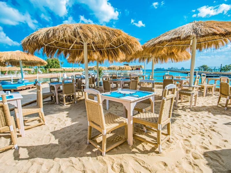Área de comedor al aire libre en la playa con sombrillas de paja y mesas en la arena.