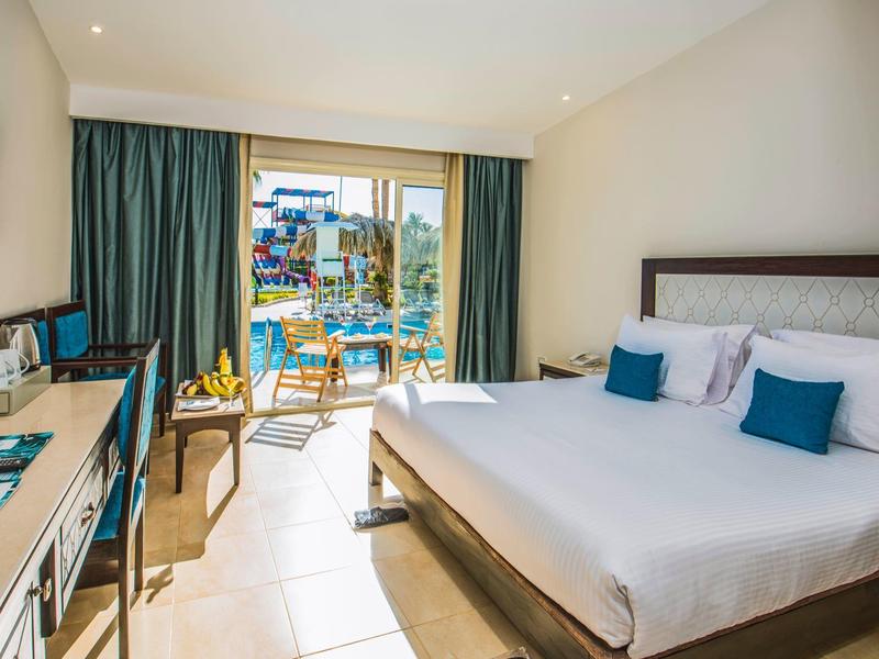 Habitación de hotel luminosa con cama doble, escritorio, balcón y vista a la zona de piscina.