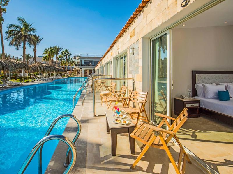Habitación de hotel con terraza junto a la piscina, equipada con muebles y palmeras al fondo.