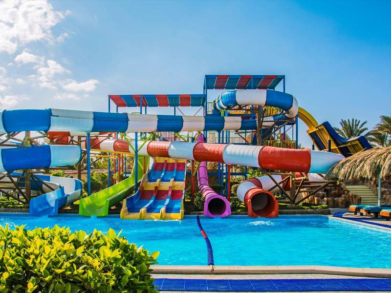 Parque acuático con toboganes coloridos, piscina rodeada de palmeras y cielo soleado.