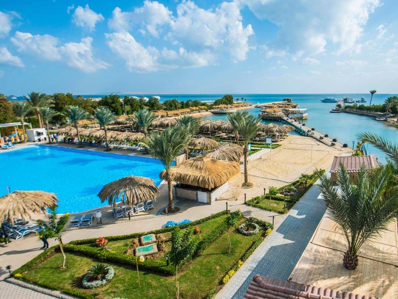 Piscina del hotel con sombrillas y palmeras, vista al mar y cielo azul con nubes.