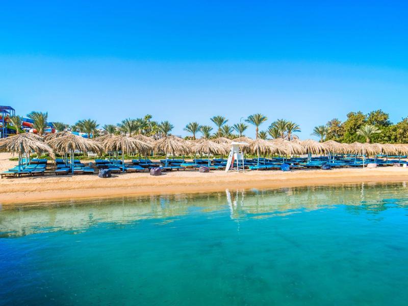 Playa de arena con tumbonas y palmeras bajo cielo azul claro y agua turquesa.