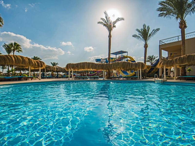 Piscina al aire libre con agua clara, sombrillas de paja y palmeras bajo un cielo azul soleado.