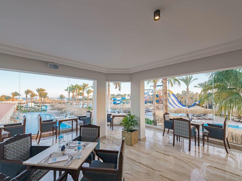 Restaurante luminoso con grandes ventanas que ofrecen vista al puerto y palmeras, mesas preparadas.