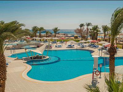 Großer Hotelpool mit Sonnenliegen und Palmen, Meer im Hintergrund.