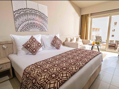 Helles Hotelzimmer mit großem Bett, gemusterten Kissen, Sofa und Balkon mit Blick nach draußen.