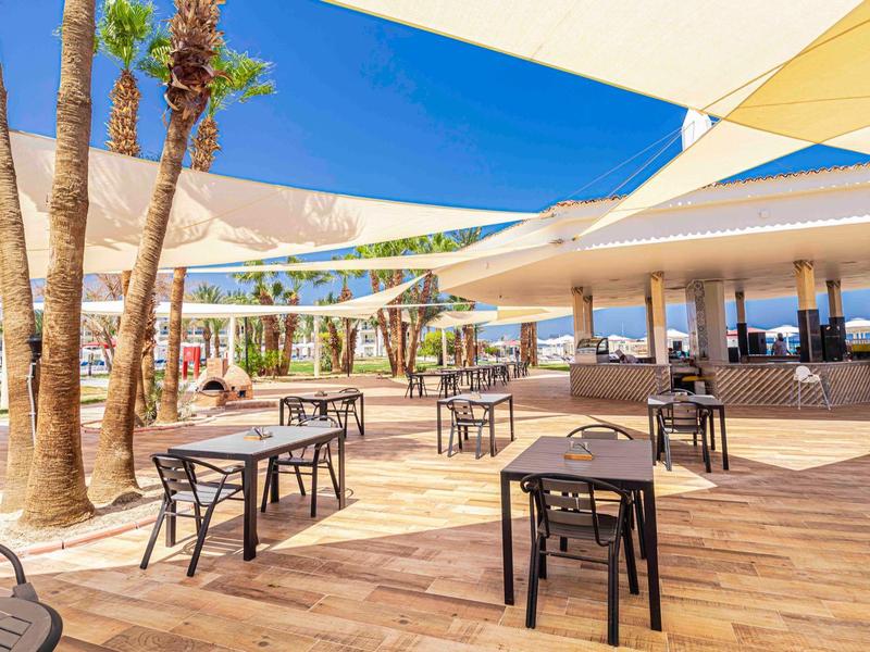 Offener Sitzbereich mit schwarzen Tischen, Sonnensegeln und Palmen auf Holzterrasse bei klarem Himmel.