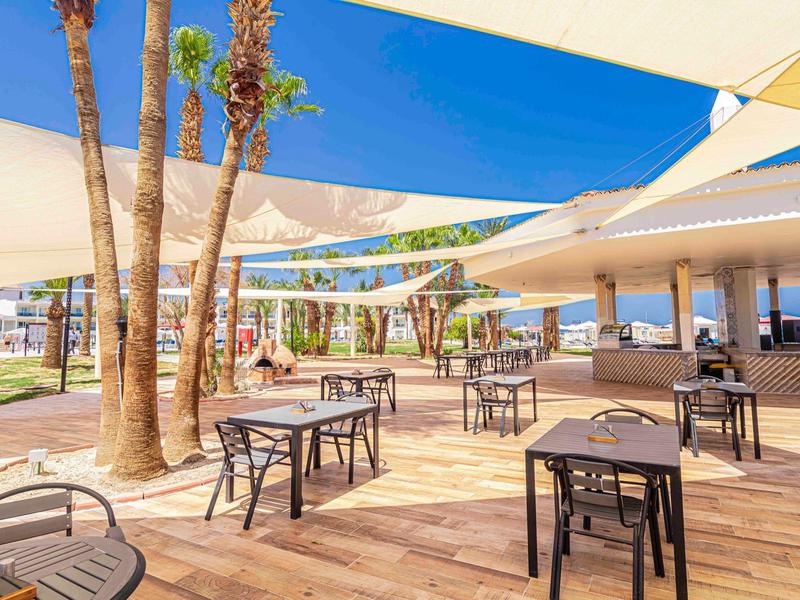 Freiluftcafé mit Holzboden, Tischen, Stühlen und großen Sonnensegeln unter blauem Himmel.