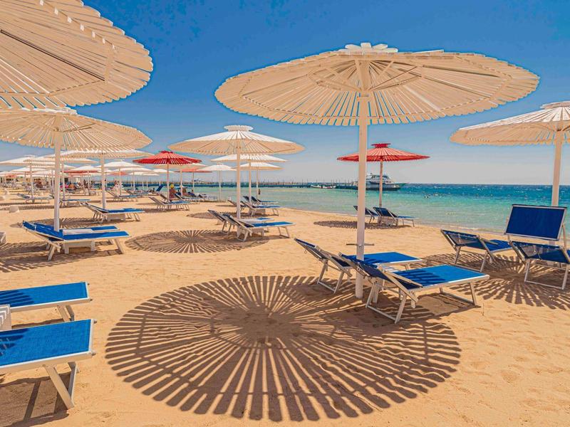 Sandstrand mit blauen Liegen und weißen Sonnenschirmen, klare blaue Meer und Himmel im Hintergrund.