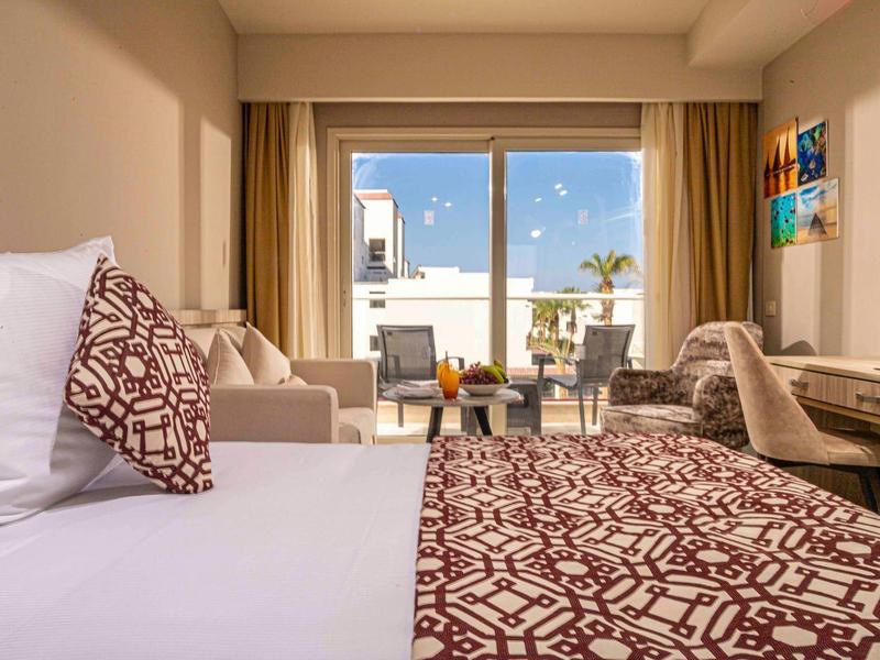 Helles Hotelzimmer mit großem Bett, gemustertem Bettüberwurf, Sitzbereich und Balkon mit Meerblick.