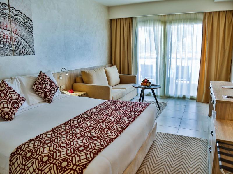 Hell eingerichtetes Hotelzimmer mit großem Bett, Sofa, Tisch, Schreibtisch und Balkon mit Vorhängen.