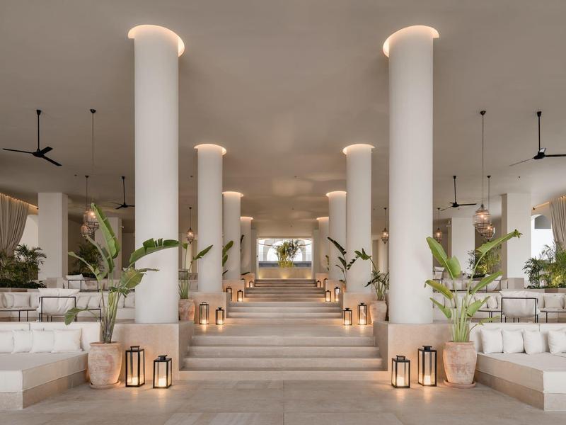Großer heller Innenraum mit Säulen, Treppe, Pflanzen und gemütlichen Sitzbereichen in sanften Farben.