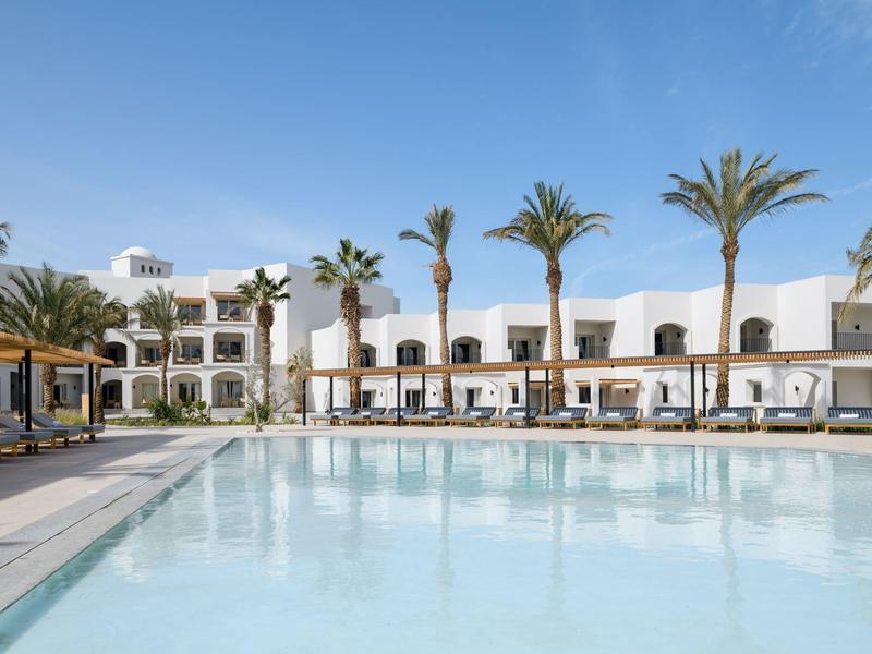 Großer klarer Pool vor weißem Hotel mit Palmen und blauem Himmel im Hintergrund.