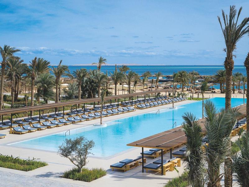 Großer blauer Pool mit vielen Liegen und Palmen, im Hintergrund das blaue Meer unter leicht bewölktem Himmel.