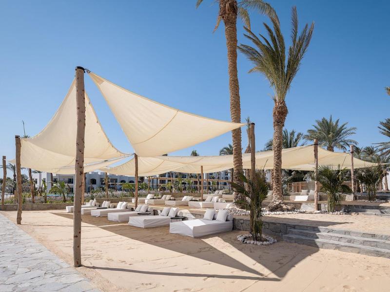 Strandbereich mit weißen Sitzkissen und Sonnensegeln unter Palmen bei blauem Himmel
