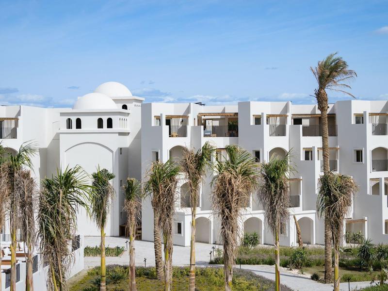 Weißes Gebäude mit Bögen, Palmen davor und blauem Himmel im Hintergrund.