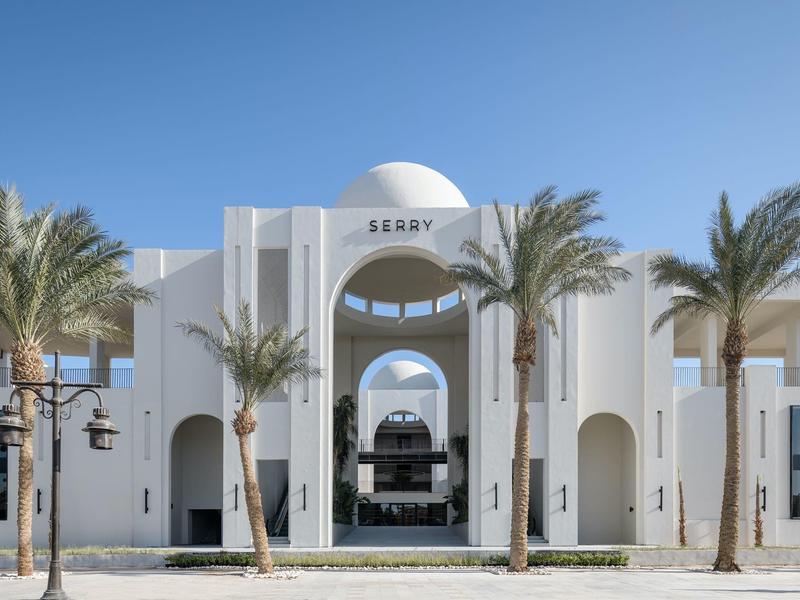 Weißes Gebäude mit großen Bögen, Kuppel und Palmen vor blauem Himmel.