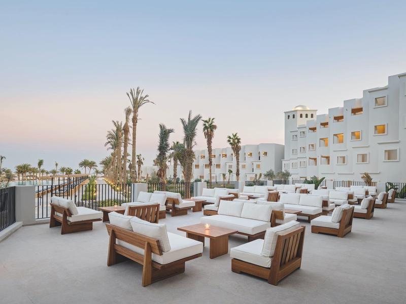 Geräumige Außenterrasse mit weißen Sofa-Sets, hölzernen Akzenten, Palmen und mehrstöckigem weißen Gebäude.
