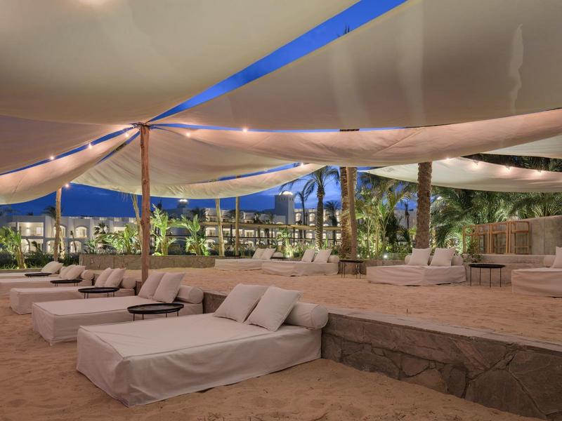 Offener Lounge-Bereich mit großen weißen Polstern, Sandboden und farbigen Segeln als Überdachung.
