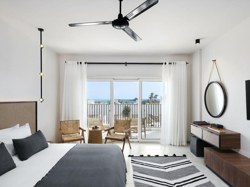 Helles Schlafzimmer mit weißem Bett, zwei Sesseln, großem Fenster und modernem Ventilator an der Decke.