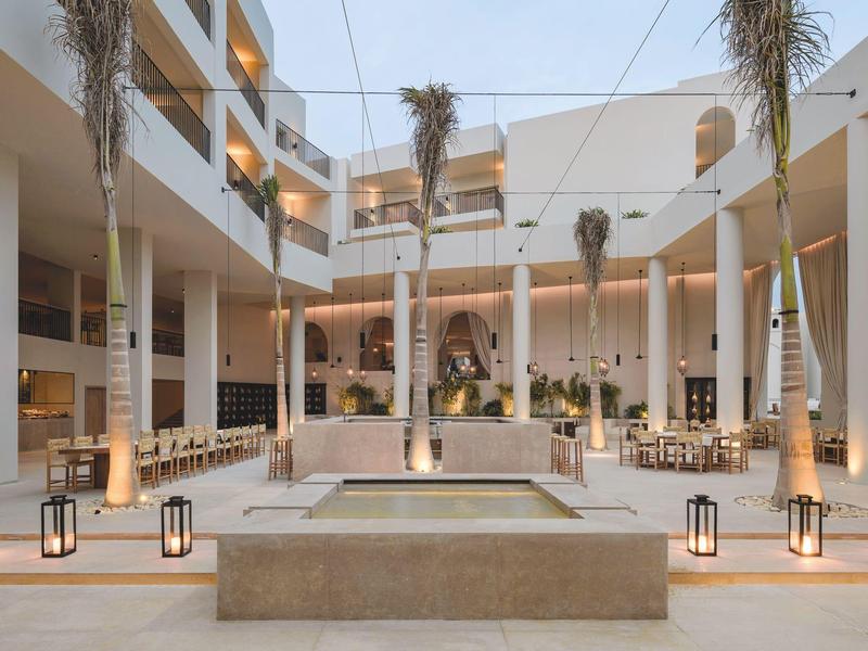 Moderner heller Innenhof mit Palmen, Säulen, Wasserbecken und stilvollen Laternen.