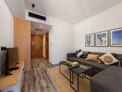 Modern eingerichtetes Hotelzimmer mit graßer Eckcouch, Fernseher und Obst auf einem kleinen Tisch.