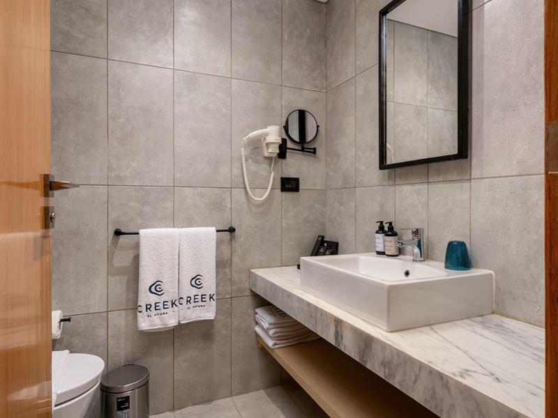 Modernes Badezimmer mit grauen Fliesen, weißen Waschbecken, Handtüchern und Haartrockner an der Wand.