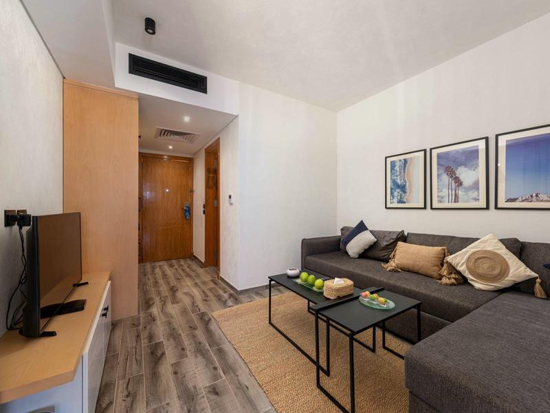 Modern eingerichtetes Hotelzimmer mit graßer Eckcouch, Fernseher und Obst auf einem kleinen Tisch.