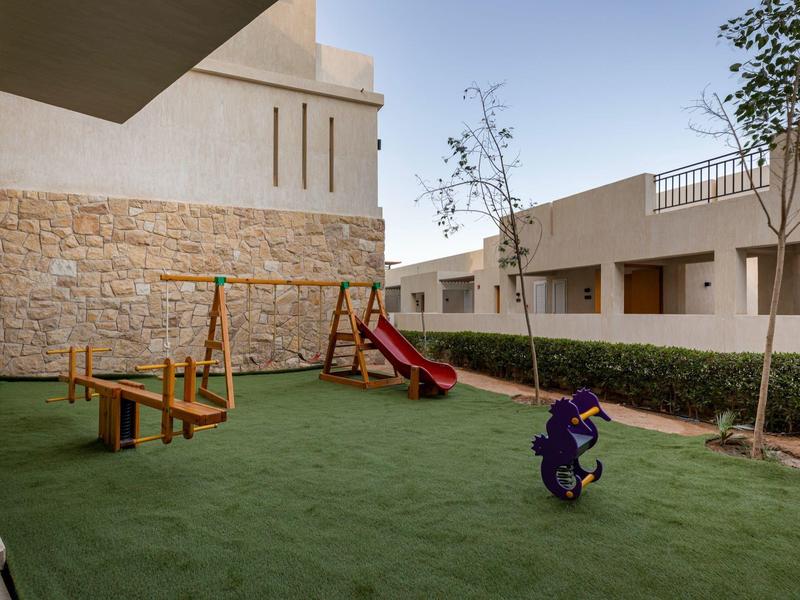 Kleiner Spielplatz mit Rutsche, Schaukeln und Wippe auf grünem Kunstrasen neben modernen Gebäuden.